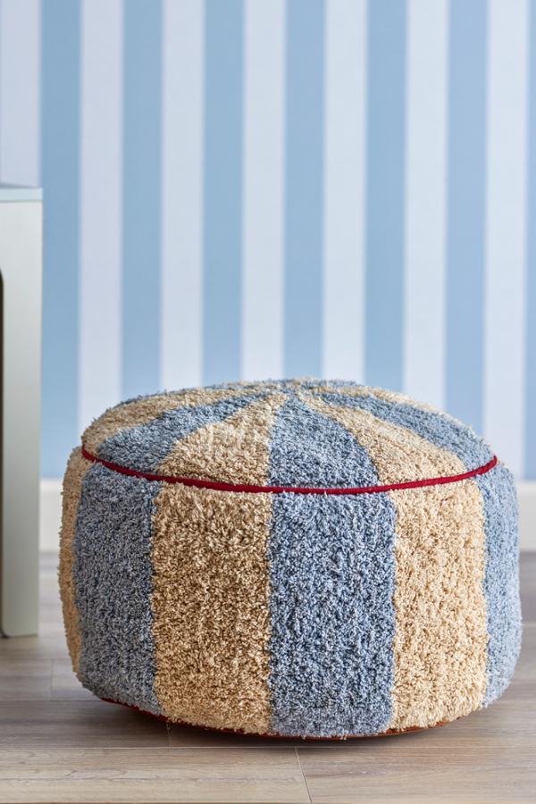 Charoline Pouf Multi – Bloomingville MINI: Pouf vor einer Wand mit blau gestreifter Tapete.